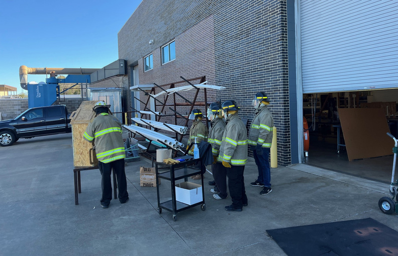 teen boys in firefighter gear watching instructor set fire to dollhouse outside building