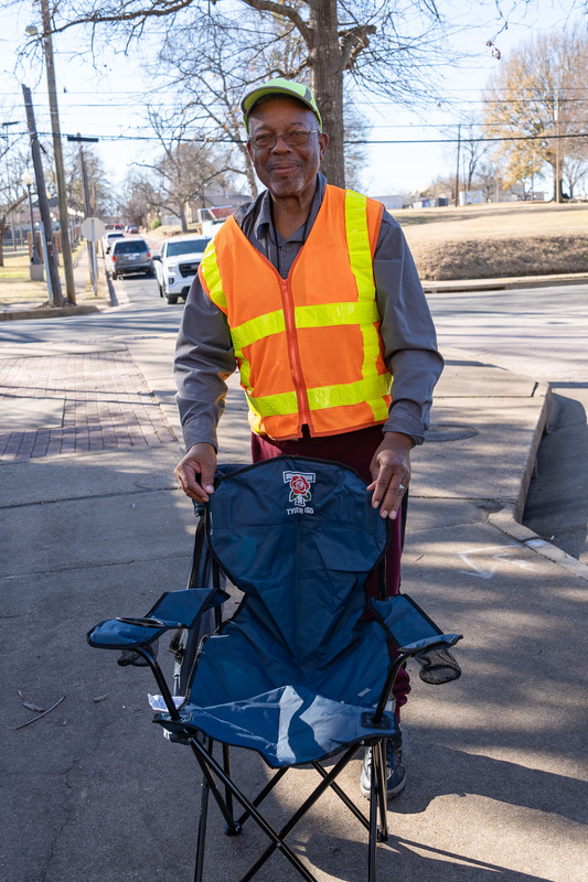 man wearing yellow vest standing behind a chair on the sidewalk