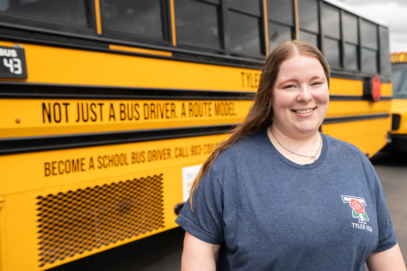 woman standing beside school bus. Side of bus says, "Not just a bus driver. A route model. Become a school bus driver, call 903-262-1000."