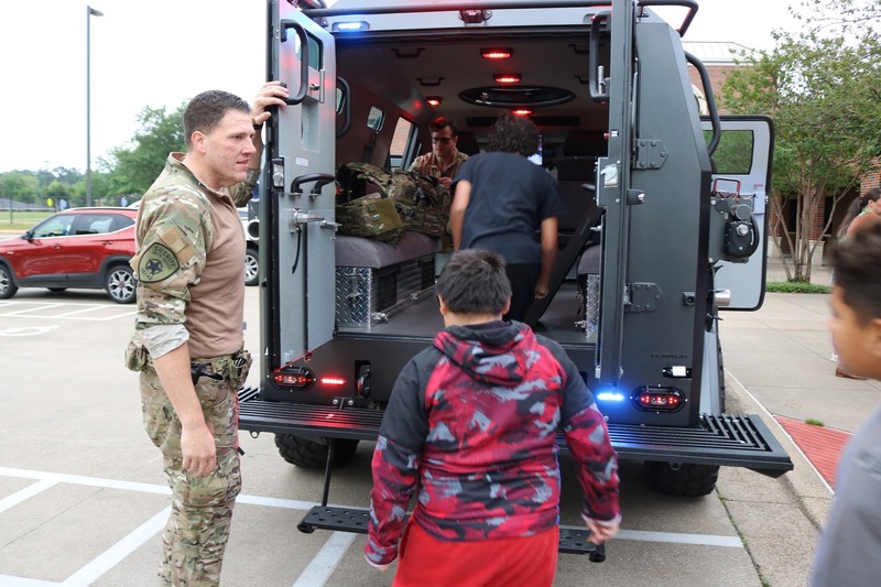 man in camo holding back door of SWAT vehicle for kids to get into