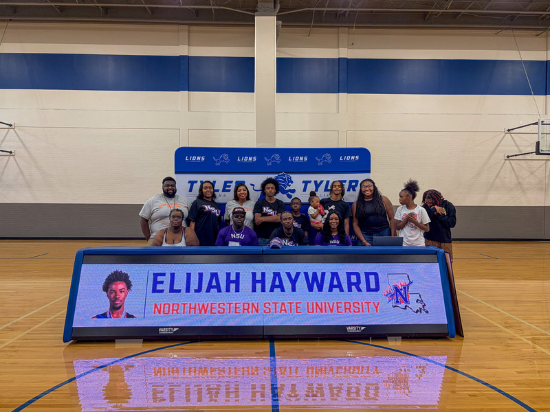 men, women, and children sitting and standing behind a table in a gym with banner on front, "Elijah Hayward, Northwestern State University."