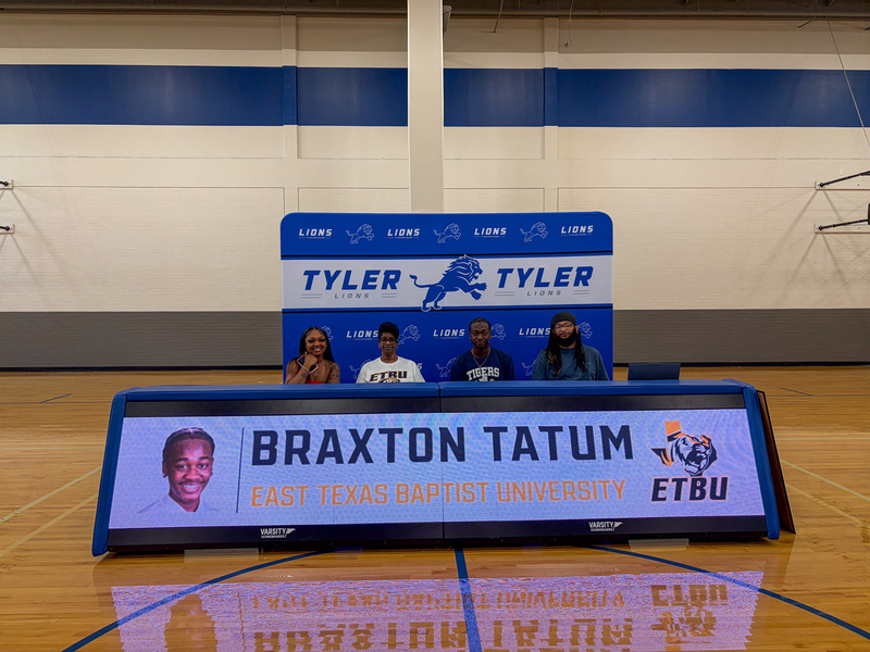 men and women sitting behind a table in gym with banner on front, "Braxton Tatum, East Texas Baptist University."