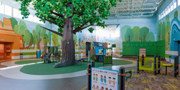 A colorful indoor children's play area featuring a large decorative tree, vibrant woodland mural, and interactive play structures.