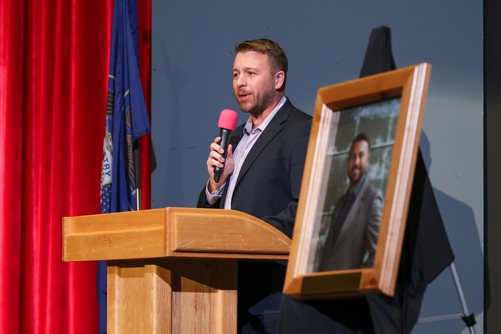 Del Dickson, Uintah High School's 2026 Distinguished Alumni, speaks at a podium during a school assembly. A framed portrait of Dickson is displayed on an easel beside him. A Utah state flag hangs in the background.