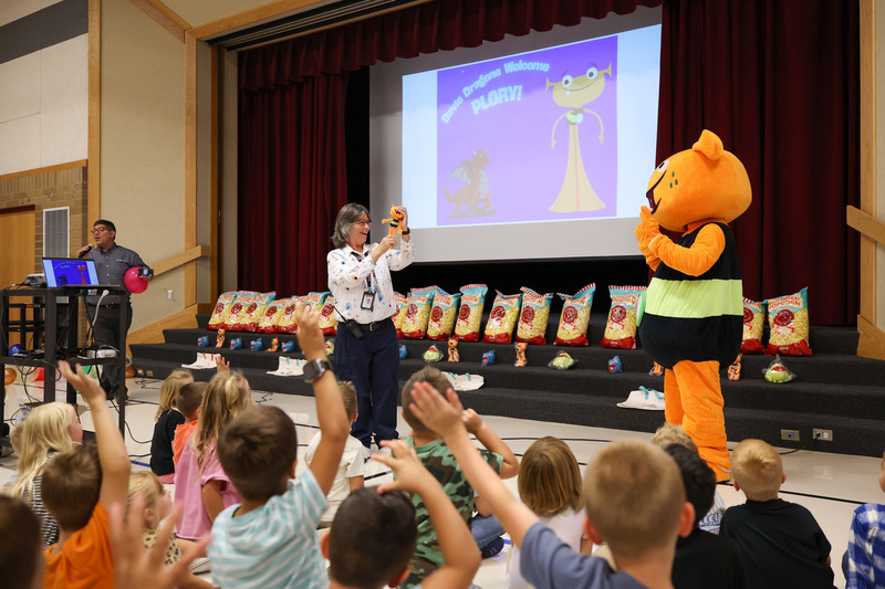 A woman holds a plush doll of an orange alien as a life-size version of the alien mascot stands to the side during an assembly in the school gym. Elementary students raise their hands in the foreground.