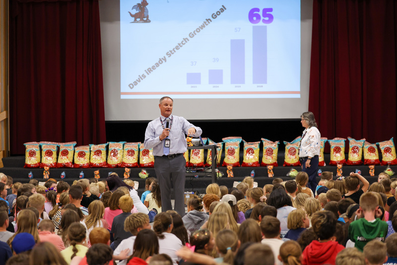 A man with a microphone stands in the middle of a group of elementary students who are seated on the floor during an assembly in the school gym.