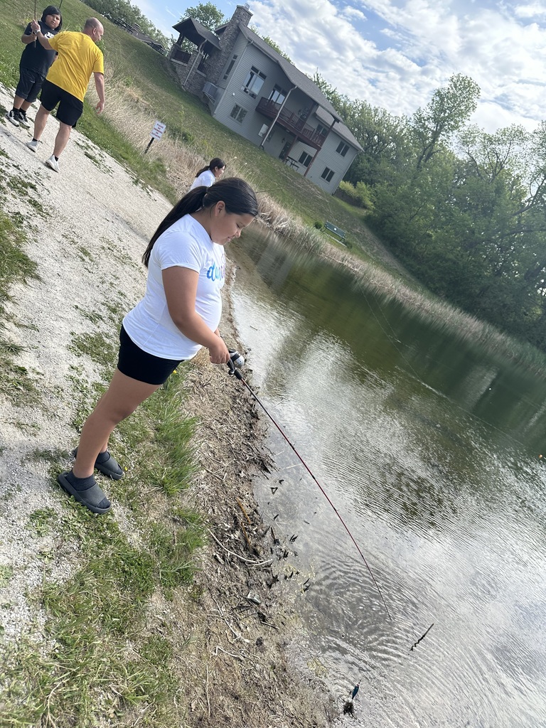 Student fishing alongisde a lake with Mr. Redden and other students in background