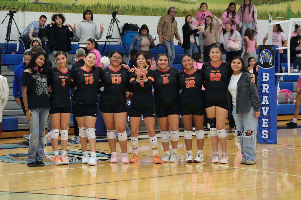 Team photo: Seven Umoⁿhoⁿ Nation volleyball players in black jerseys with red-and-gold trim (numbers 1, 15, 13, 4, 5, 12 and 26) stand arm in arm on the court, smiling. Two people in street clothes stand at either end. Number 4 makes a hand sign at the center. Fans in pink fill the bleachers behind them, next to a blue “Braves” net post.