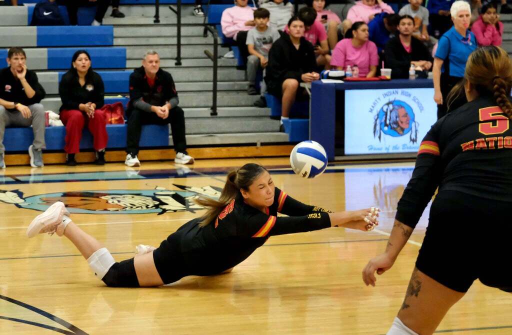 Diving dig: A Nation player dives fully stretched across the floor to keep the ball alive with her taped hands, eyes squeezed shut. Teammate number 5 is close by. Coaches watch from the bench next to the Marty Indian High School scorer’s table.