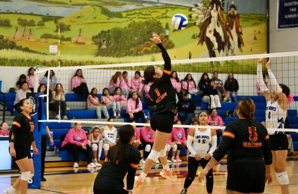 Attack over the block: Nation’s number 1 leaps high to hit the ball as Lady Braves number 4 jumps to block. Nation’s numbers 15 and 5 and a crouching teammate cover the play. A painted mural of riders on horseback and a crowd in pink shirts fill the background.