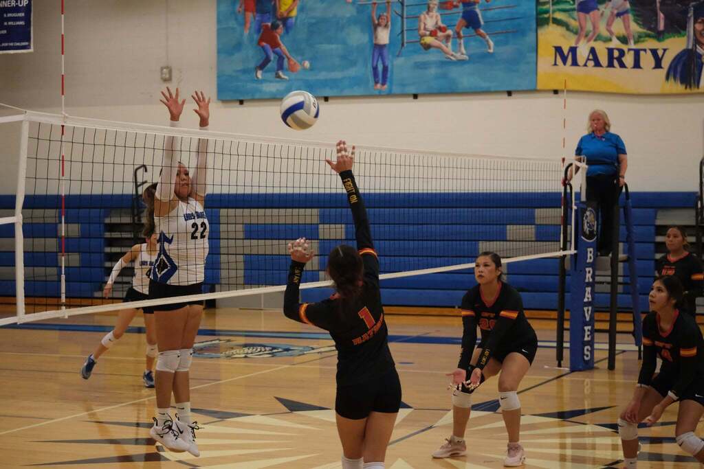 Set at the net: Umoⁿhoⁿ Nation’s number 1 reaches up to set the ball while a Lady Braves player in white, number 22, jumps to block. Two Nation teammates crouch, ready. A referee watches from the stand under a colorful “Marty” mural.