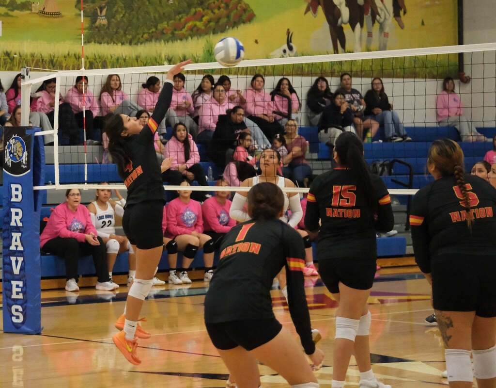 Jump at the net: A Nation player jumps beside the blue “Braves” post to play the ball at the net. Teammates 1, 15 and 5, seen from behind, watch the play. The Lady Braves bench and a crowd in pink sit beyond the net.