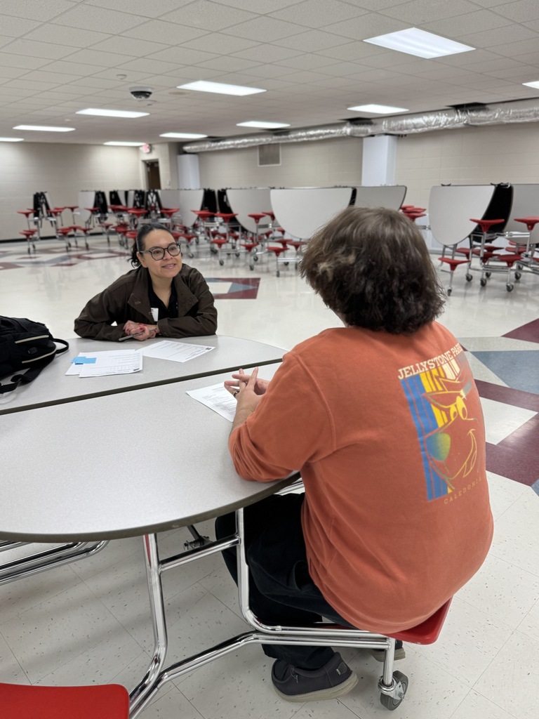 Two people sit at a table in an empty room with red chairs and white walls.