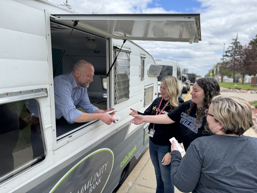 A man serves food to three women standing outside a food truck with a road and trees in the background.