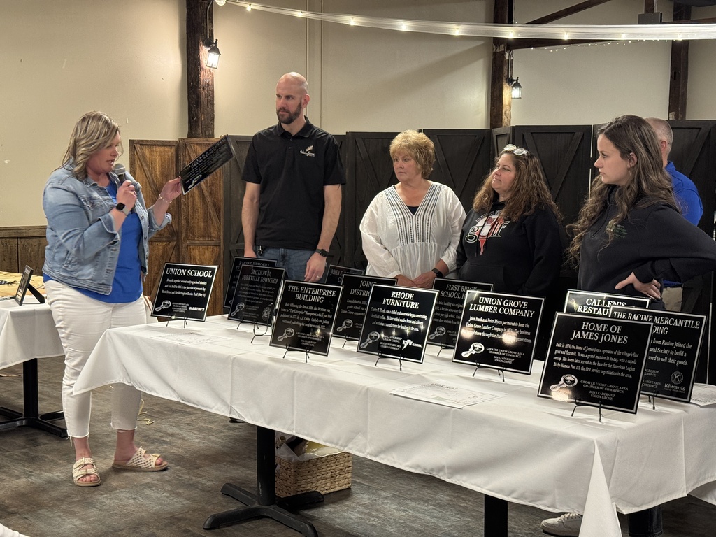 A group of people stand around a table with awards on it. A woman holds a microphone.