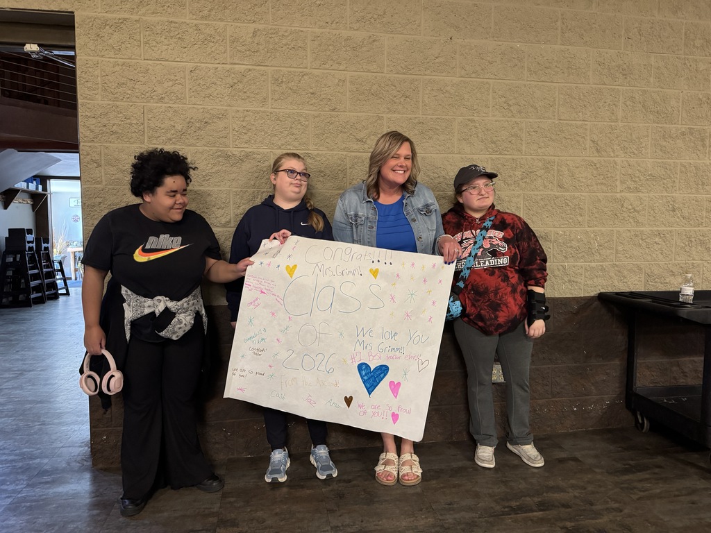 Four people standing, one holding a large white sign with hearts and text, against a beige wall.