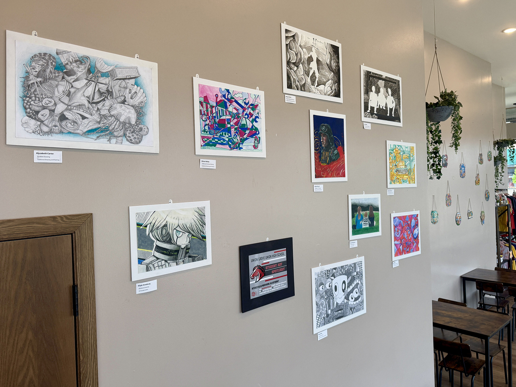 Multiple artworks hang on a wall, featuring diverse styles and colors. A wooden door and tables with chairs are visible.