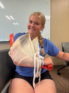 A person with a bandaged arm smiles while sitting in a chair. Behind them is a whiteboard.