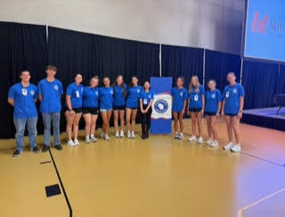 A group of athletes in blue shirts and shorts pose together on a court with a logo.