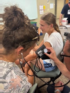 A person measures a woman's blood pressure. Both sit in chairs. The woman wears a white shirt.