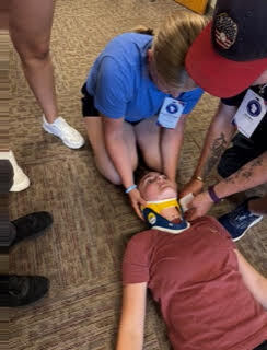 An individual lies on the ground wearing a neck brace. Two people kneel to stabilize the person.