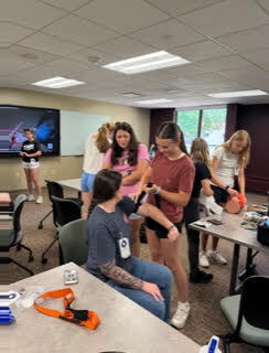 Group of people inside a classroom, one person sitting and others standing. One person taking blood pressure.