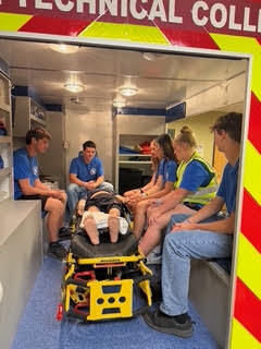 A group of people in blue shirts and safety vests sit in a mock ambulance. A person lies on a stretcher.