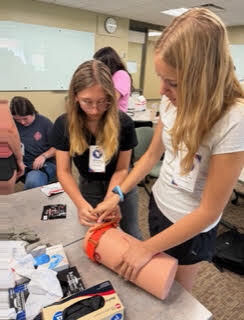 Two individuals, one seated and another standing, working on a training dummy's arm. Various medical items on a table.