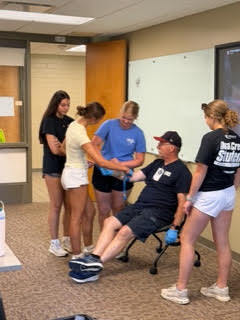 A group of five people in a classroom, one seated on a chair receiving blood pressure check.