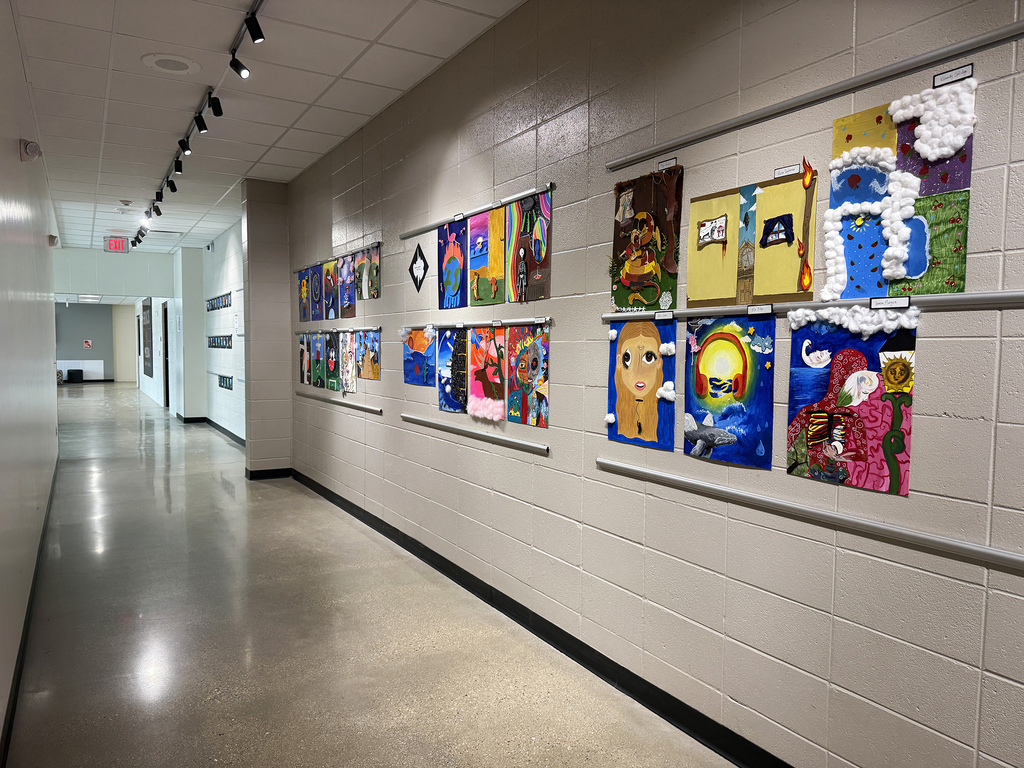 A long, empty hallway with white walls. Colorful art pieces are displayed on one side wall, in rows.