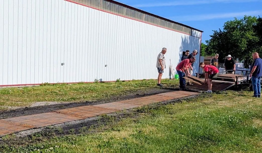 People work on a new pathway in front of a building. They are surrounded by grass and trees.