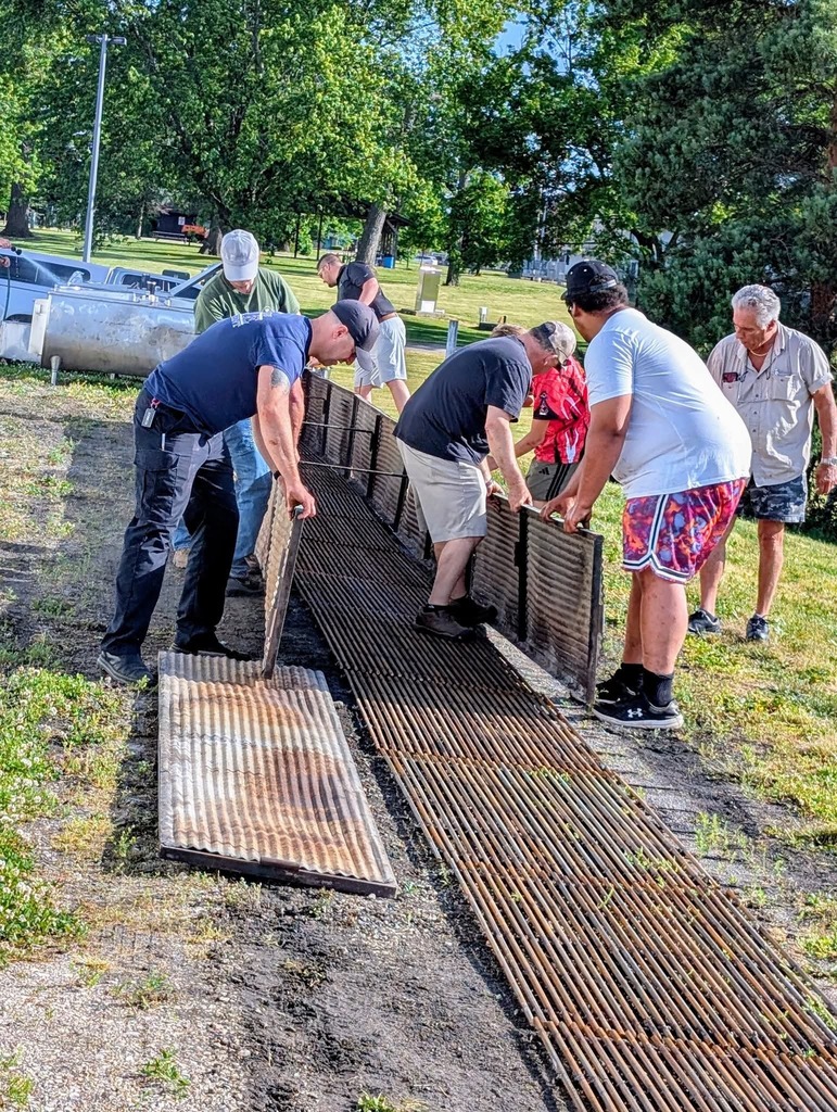 Several people are installing a wooden structure in a grassy area. They wear casual clothing. Trees and a vehicle are in the background.