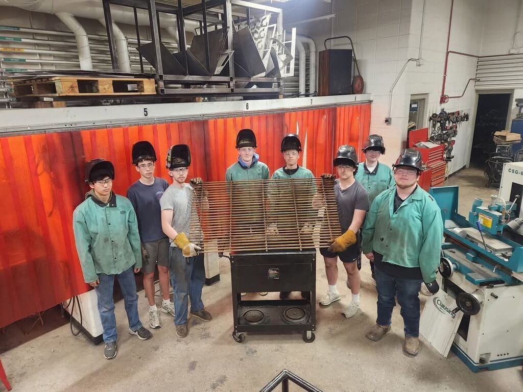 Nine people wear safety gear. They hold a metal screen in front of a red curtain.