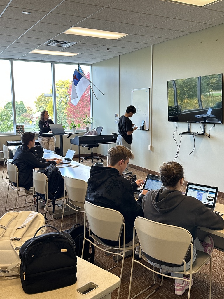 three students working on chromebooks and one drawing on the whiteboard while the teacher watches
