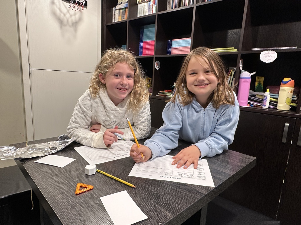 two girls working on an assignment together writing on paper and smiling 