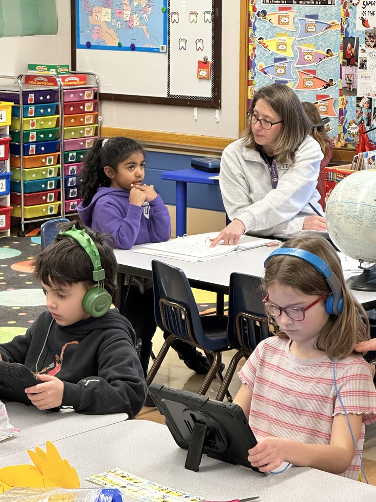 kindergarten students working on their iPads with a teacher and student working one-on-one in the background
