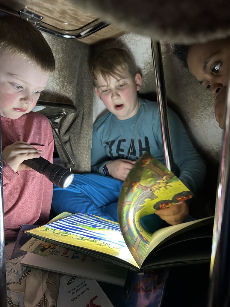 5th grade and kindergarten students reading books together by flashlight