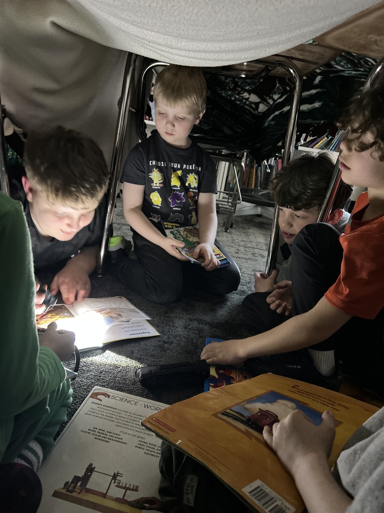 5th grade and kindergarten students reading books together by flashlight