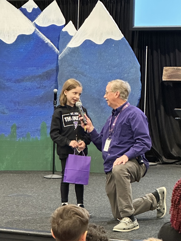 principal kneeling down next to 1st grade student holding a microphone for her to talk