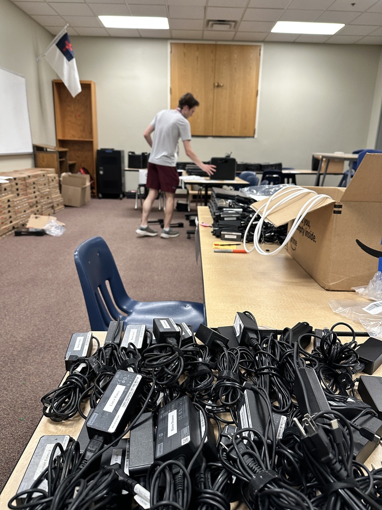 pile of device chargers on a table and a man holding a chromebook in the distance
