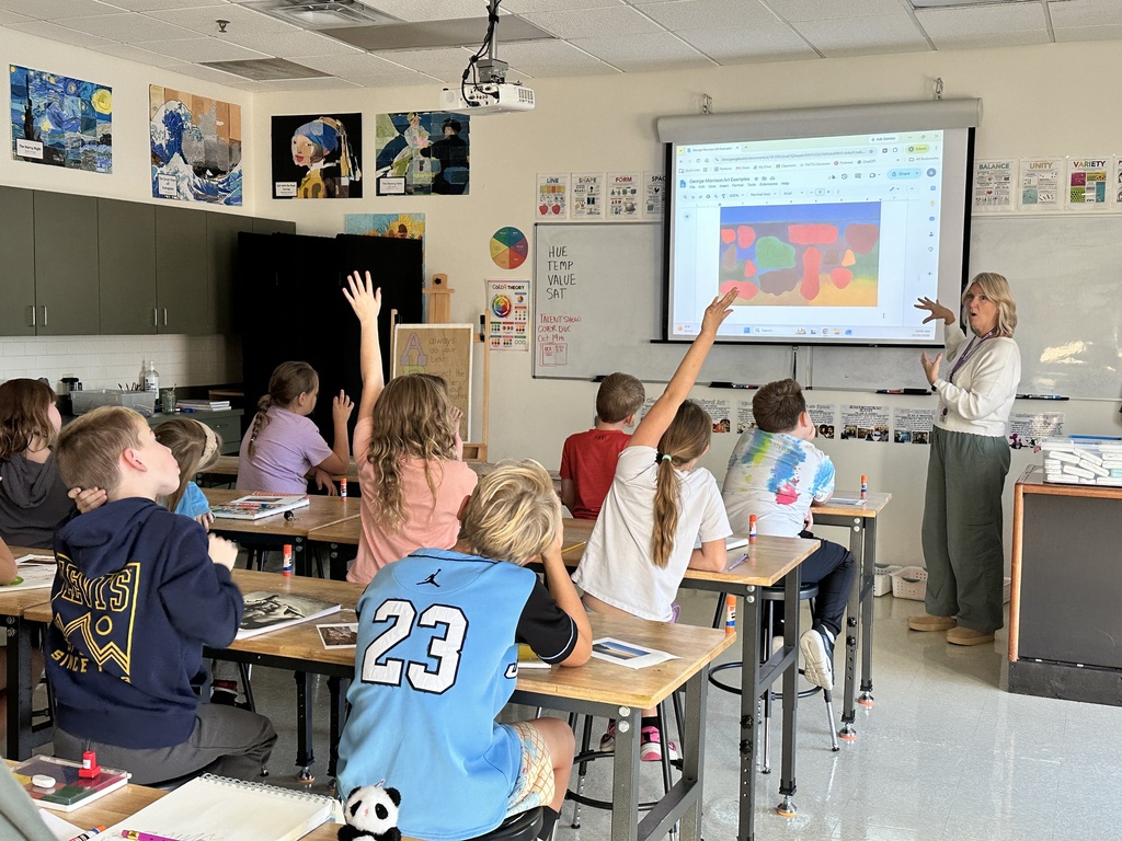 teacher in the front of the room at the white board and students raising their hands at their desks