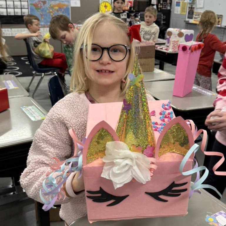 Smiling child in a classroom holding a pink, glittery unicorn-themed Valentine’s box.