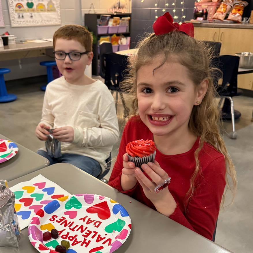 A student in a red shirt holding a Valentine's Day cupcake at her desk