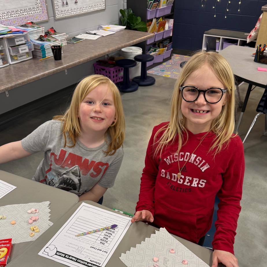 Two students smiling at their desk while completing a candy heart math worksheet