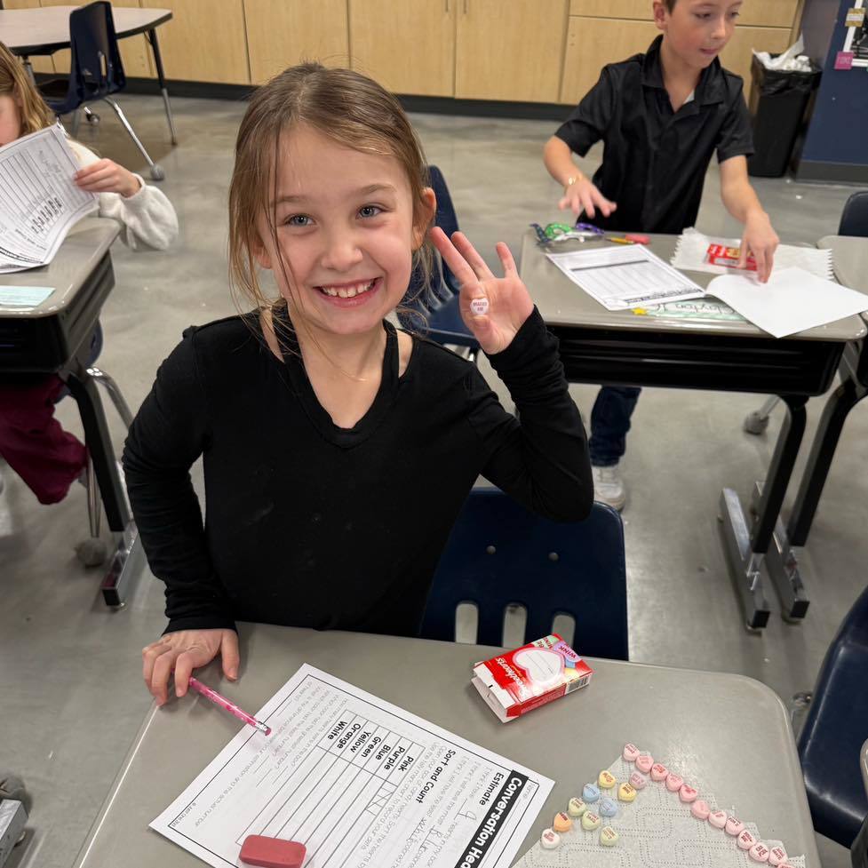 For the photo of the student at her desk, here is a concise alt text:  "A smiling student at her desk working on a Valentine's Day candy heart math activity