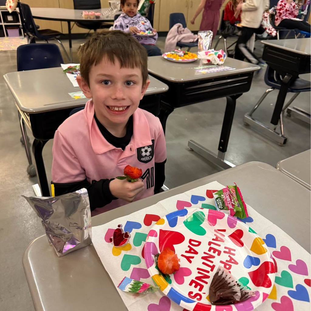 picture of A student in a pink polo smiling at his desk with a heart-themed Valentine's Day project