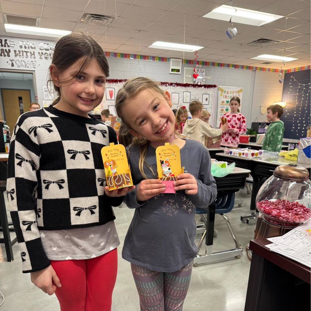 Two smiling students standing together in their classroom holding their Valentine's Day cards