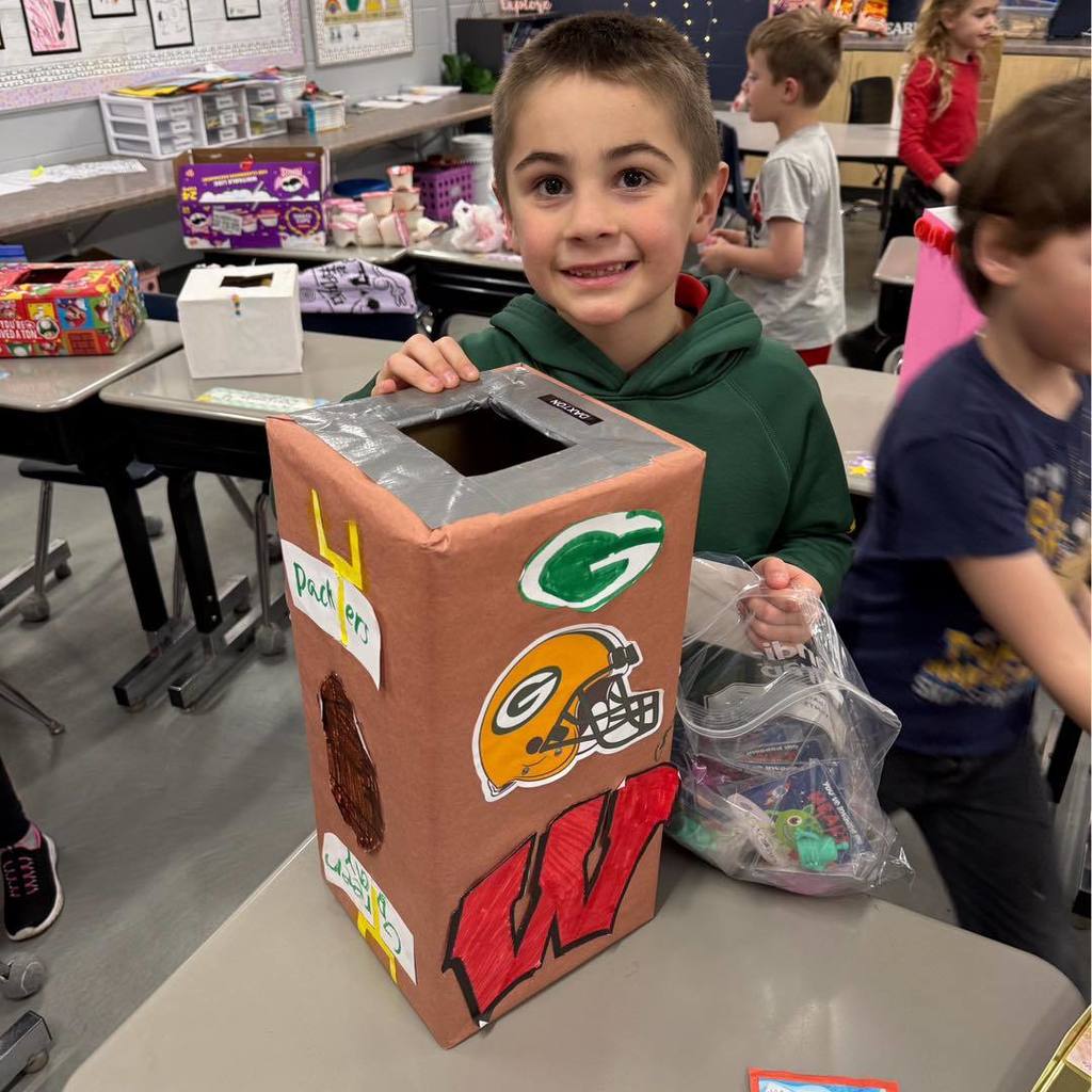 A student in a green hoodie smiling with his Valentine's Day box decorated with Green Bay Packers and Wisconsin Badgers logos