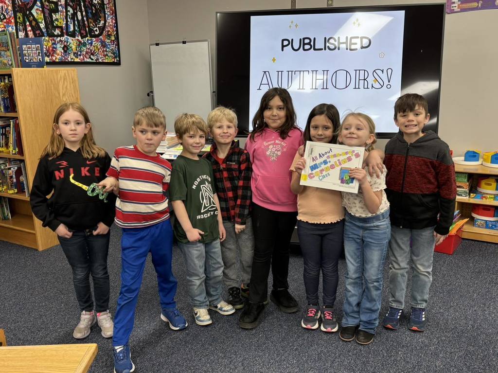 a picture of A group of seven elementary students standing in a classroom, with one student holding a book titled "All About Mrs. Cornelius's Class." A television screen in the background displays the words "PUBLISHED AUTHORS!" in blue and black text