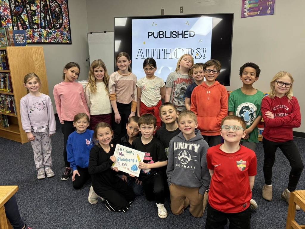 picture of A group of 15 elementary students posing together in a classroom. One student in the front holds a book titled "All About Ms. Blomberg's Class," while a television screen in the background displays the message "PUBLISHED AUTHORS!